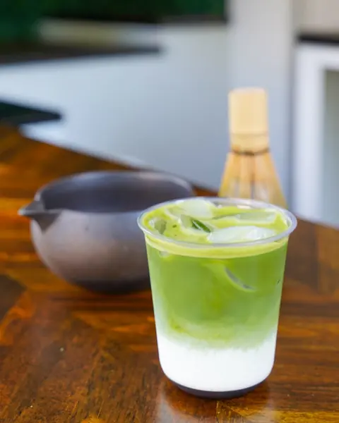Matcha helado clásico sobre leche de avena en un vaso transparente, con un tazón de matcha y un batidor de bambú detrás.